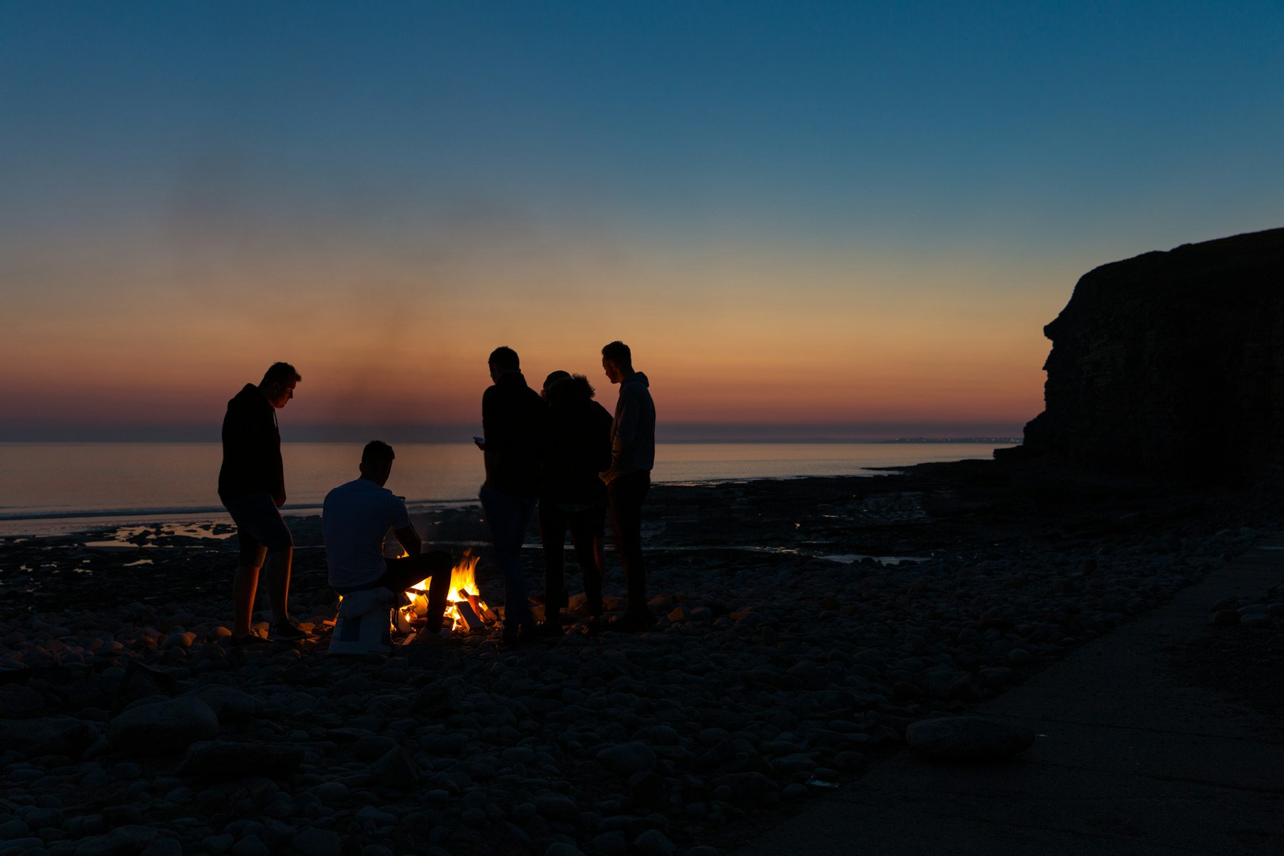 Silhouettes of people gathered around a bonfire by the beach at sunset