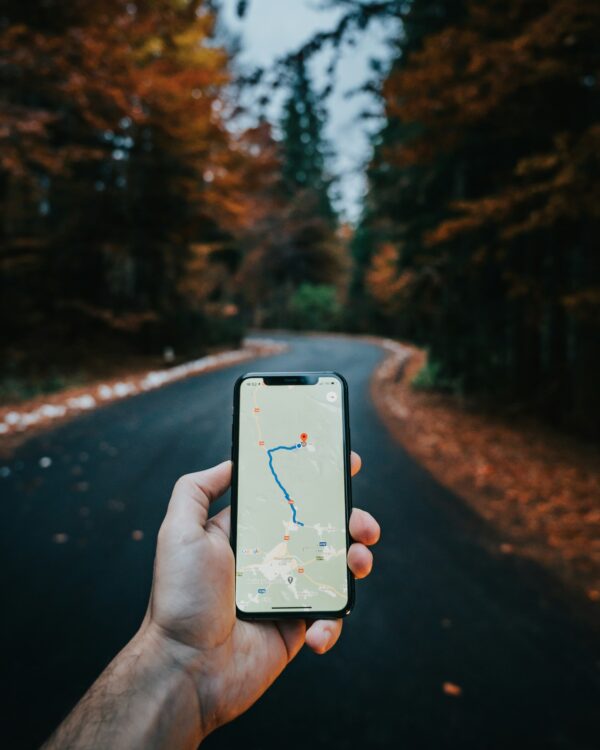 A hand holding a smartphone showing a route map on a tree-lined road. Photo by Tamas Tuzes-Katai on Unsplash.